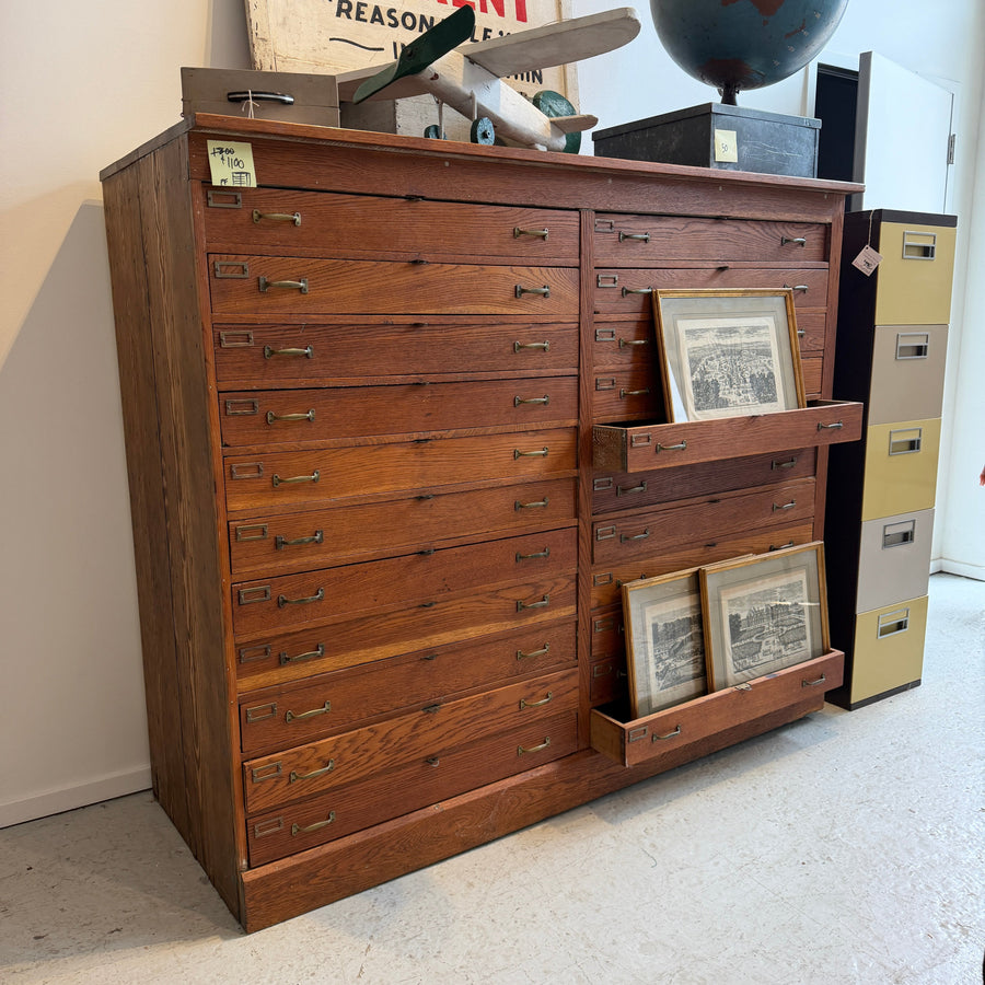 Vintage wooden multi-drawer flat file cabinet with shallow drawers and brass-tone pulls, with framed prints displayed in trays.